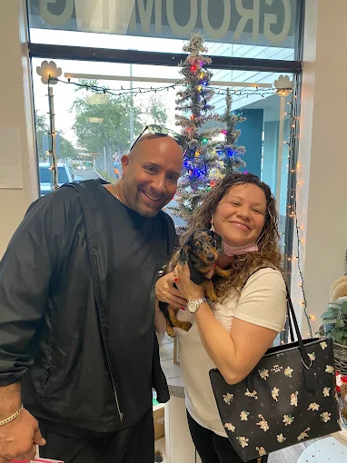 Two people holding a small puppy inside Wags About You
